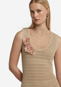 Beige virkad linne med rundad halsringning, med tre lager av rosa blommönster applicerade på vänster axel.