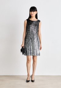 Abito in paillettes nere e argento con scollatura rotonda e maniche corte, caratterizzato da un design svasato. Modella che tiene una pochette nera e indossa tacchi.