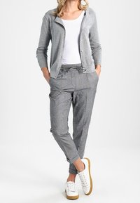 Kvinna som bär en grå cardigan, vit t-shirt, grå rutiga dragsko-byxor och vita sneakers med gummisulor, står med händerna i fickorna.