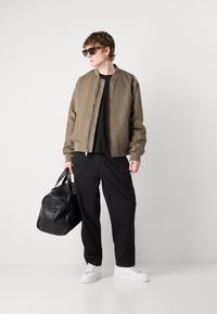 Veste bomber beige, t-shirt noir, pantalon large noir, lunettes de soleil noires et baskets blanches. Tissu lisse, coupe décontractée, design minimaliste.
