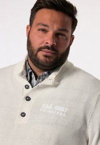 Elfenbeinfarbenes Strickpullover mit einem gerippten Kragen, drei dunklen Knöpfen und dem weißen Text "SAIL AWAY". Wird über einem karierten Hemd in Blau- und Grautönen getragen.