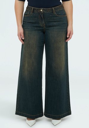 Vidbenede mørkeblå jeans med falmede lår og små kobberfarvede nitter, båret med hvide spidse sko.