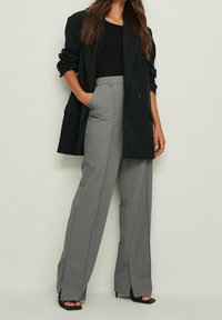 Graue maßgeschneiderte weite Hose mit Schlitz vorne, kombiniert mit einem schwarzen enganliegenden Oberteil und einem langen schwarzen Blazer mit einem einzelnen Knopfverschluss.