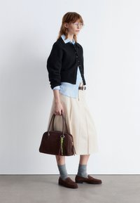 Schwarze Strickjacke, hellblauses Hemd, beigefarbener Plissee-Rock, braune Wildlederhandtasche mit buntem Anhänger, graue Socken und braune Slipper.