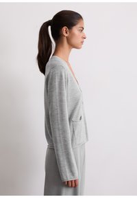 Cardigan grigio chiaro realizzato in tessuto morbido, con maniche lunghe, scollo a V e due tasche frontali, progettato con una vestibilità comoda.
