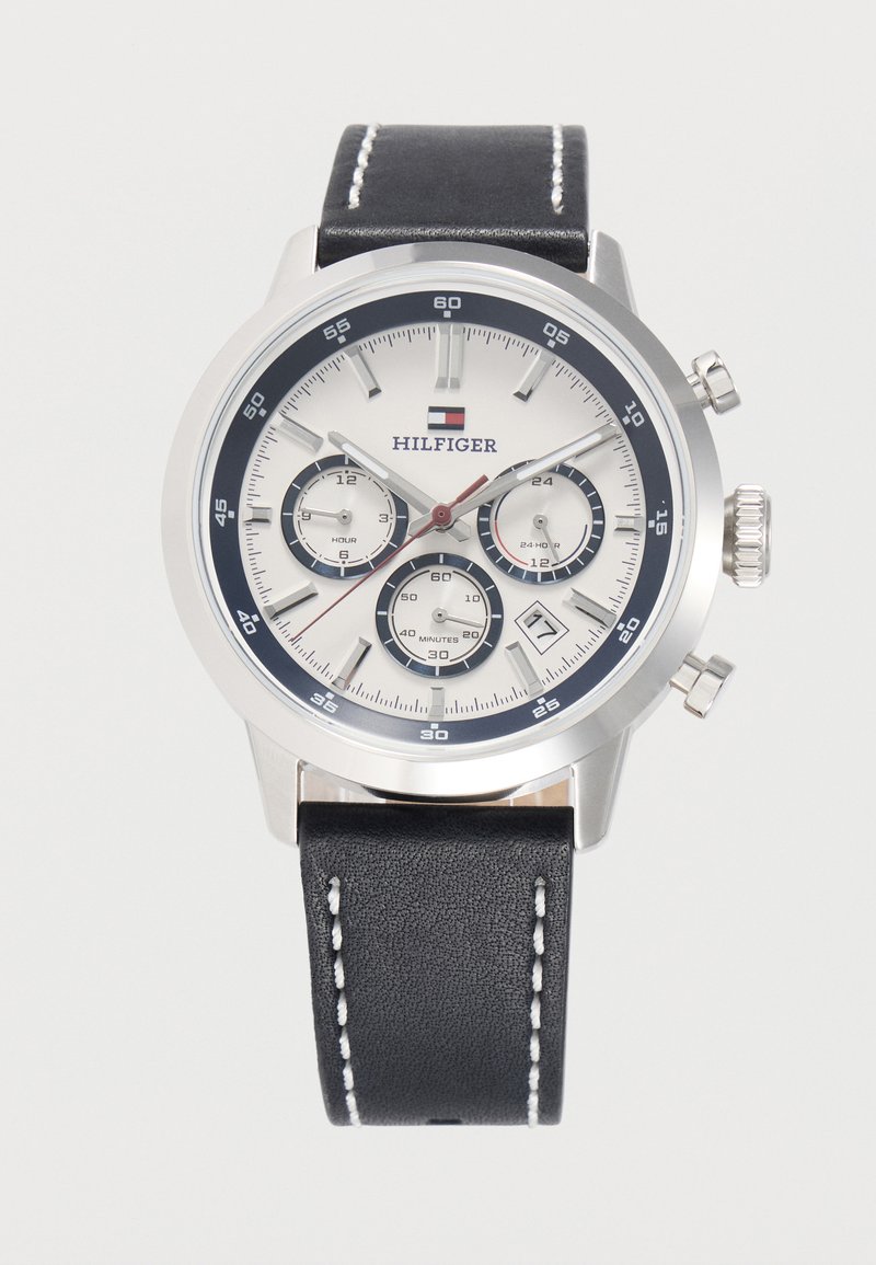 Στρογγυλό ασημένιο ρολόι χειρός με λευκό καντράν, μαύρο δερμάτινο λουράκι με λευκό ράψιμο, τρεις υποκαντράν, παράθυρο ημερομηνίας στη θέση 4 και λογότυπο της μάρκας.