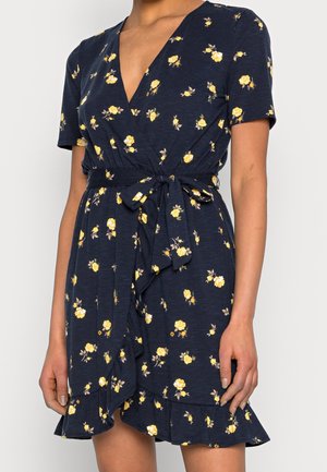 Short-sleeve navy dress with yellow floral pattern, V-neckline, tie waist, and ruffled hem worn by standing person.