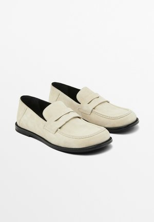 Beige suède instappers met een platte zwarte zool, voorzien van een gladde neus en decoratieve stiksels op de bovenzijde. Eenvoudig en klassiek ontwerp.
