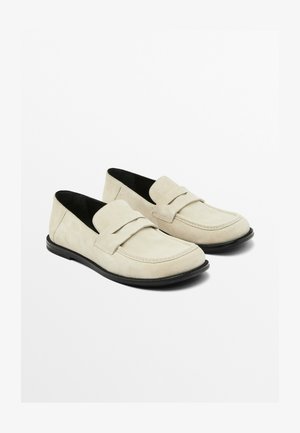 Beige ruskind loafers med flad sort sål, med en glat tå og dekorativ syning på vampen. Enkel og klassisk design.