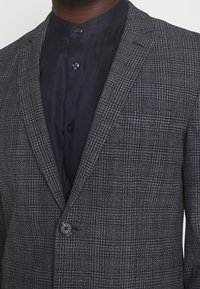 Sivý blejzr s kostkovaným vzorem a matnou texturou, se zapínáním na jeden knoflík a klopami, přes tmavou košili na knoflíky.