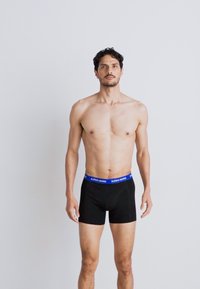Fekete boxer alsónadrág kék márkázott derékpánttal, testhezálló fazon, sima anyag és rövid szárhossz, egy álló férfi által modellezve.