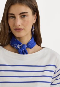 Écharpe à motif floral bleu nouée autour du cou, assortie à une chemise blanche à rayures horizontales bleues. Les cheveux sont longs et foncés.
