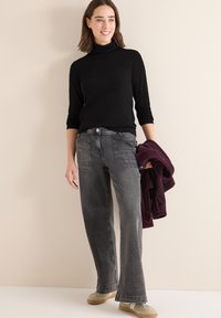 Czarny golf, szare szerokie jeansy i beżowe sneakersy. Trzyma burgundowy sweter z dzianiny. Gładki materiał, casualowy wygląd. Minimalistyczny design.