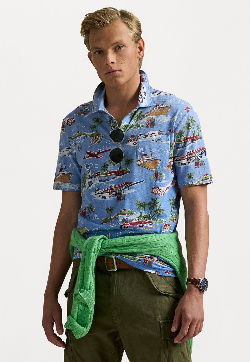 Korte mouwen blauwe shirt met een tropische print van boten en palmbomen, gecombineerd met een groene tricot rond de taille en beige cargobroek.