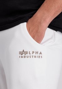 Witte sweatpants met een voorzak, voorzien van een tan geborduurd logo met de tekst "ALPHA INDUSTRIES." Zacht, glad katoenweefsel.