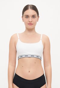 Bralette bianca con spalline sottili e una fascia nera con logo. Texture liscia, design senza imbottitura e uno stile minimalista e sportivo. Sotto, indossa della biancheria intima nera.