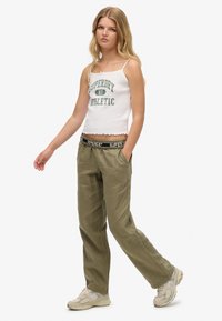 Débardeur blanc court avec imprime vert ; pantalon ample olive avec une ceinture de marque noir et blanc ; baskets dans des tons neutres.