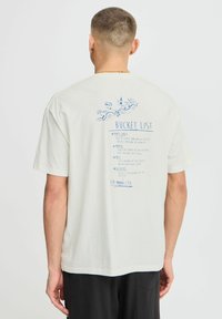 Light-colored cotton t-shirt featuring a blue printed "Bucket List" design on the back, including illustrations and text details.