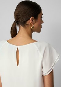 Witte blouse met korte, zwevende mouwen en een sleutelgatdetail op de achterkant, vastgemaakt met een klein knoopje. Soepele, lichte stof.