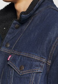 Denim jacket in dark blue with a shearling collar, featuring metal buttons, contrast stitching, and a chest pocket with logo label.
