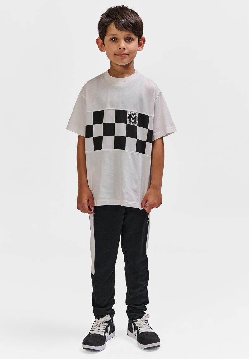 Junger Junge steht mit dem Gesicht nach vorne und trägt ein weißes T-Shirt mit schwarzem Karomuster, schwarze Jogginghosen mit weißen Streifen und schwarze Turnschuhe.