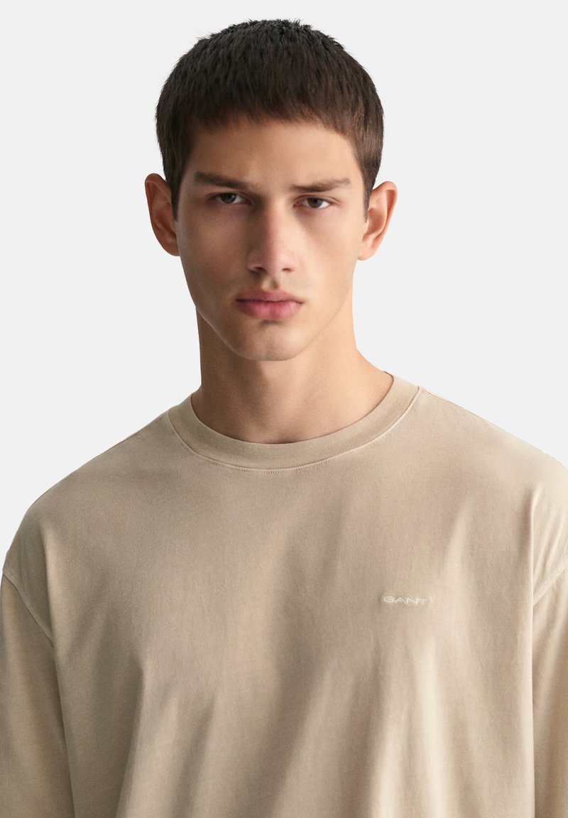 Camiseta de algodón beige con cuello redondo, hombros caídos y un sutil logo bordado en el pecho. Textura suave, corte relajado.