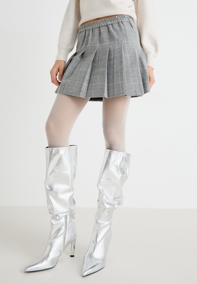 Personne portant une mini-jupe plissée grise, des collants blancs et des bottes argentées brillantes à talons hauts jusqu’aux genoux, debout devant un fond uni.
