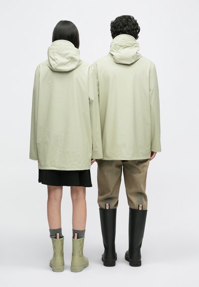 Dwie osoby stoją plecami do siebie, ubrani w jasnobeżowe kurtki z kapturem; jedna w czarnej spódnicy i zielonych butach, a druga w brązowych spodniach i czarnych butach.