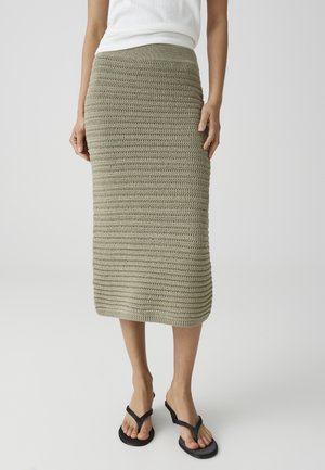 Vrouw draagt een olijfgroene gehaakte rok met hoge taille tot op de knie, zwarte flipflops met hakken en een witte top, staand op een witte vloer.