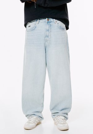 Person, der hellblaue, weite Jeans, weiße Turnschuhe und ein dunkles Langarmoberteil trägt, steht vor einem weißen Hintergrund.