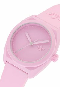 Rozā silikona pulkstenis ar apaļu ciparnīcu, atbilstošām rokām, minimālistiskiem stundu marķieriem un logo akcentu uz sejas.