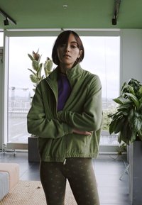 Grøn zip-up jakke lavet af blødt stof, lagdelt over en lilla top. Modellen bærer mønstrede leggings og står foran en baggrund fyldt med planter.
