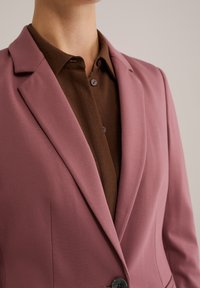 Rosa Blazer mit Revers und einem einzigen Knopf, über einem braunen Hemd mit Kragen getragen. Der Blazer hat eine glatte Textur und eine maßgeschneiderte Passform.