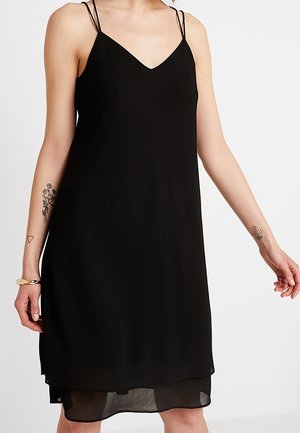 Robe noire sans manches avec double bretelles fines, décolleté en V et ourlet transparent superposé, portée par une personne avec des tatouages floraux et de mains sur les bras.