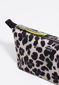 Kosmetyczka w cętkowany wzór, o stożkowatym kształcie, wykonana z materiału. Posiada czarny zamek błyskawiczny z teksturowanym zielonym uchwytem oraz detalem logo.