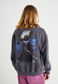 Felpa grigia scura con un grande motivo raffigurante un personaggio in una scena spaziale sul retro, con scollatura tonda e polsini e orlo a costine.