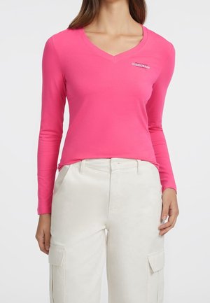 Femme portant un haut rose vif à manches longues avec col en V et logo « Guess Jeans » ainsi qu’un pantalon cargo écru avec poches avant.