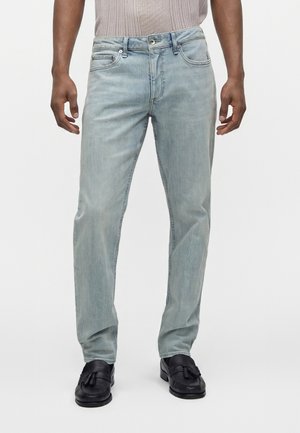 Uomo che indossa jeans azzurri chiari, maglia beige lavorata a maglia e mocassini neri con nappine, in piedi davanti a uno sfondo bianco uniforme.
