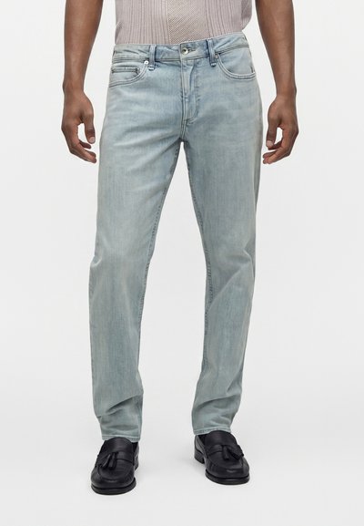 Homme portant un jean bleu clair, un haut en tricot beige et des mocassins noirs à pompons, debout devant un fond blanc uni.