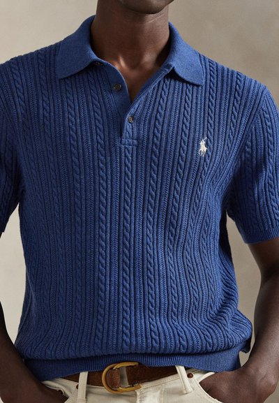 Niebieski dziergany polo z teksturowanym wzorem warkocza, krótkimi rękawami i rozcięciem na dwa guziki. Na piersi znajduje się małe haftowane logo.