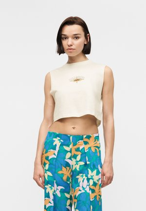 Młoda kobieta o krótkich brązowych włosach nosi bezrękawnikowy, beżowy krótki top oraz szerokie spodnie z wysokim stanem ozdobione niebieskim, zielonym i pomarańczowym wzorem kwiatowym.