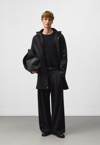 Manteau imperméable noir avec capuche, associé à un t-shirt noir et un pantalon large. Le modèle tient un sac noir. Fond de studio.