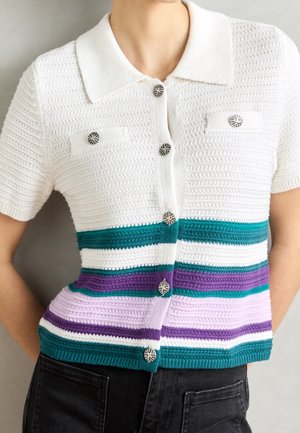 Haut en maille blanc à manches courtes avec col, boutons à motif, et rayures horizontales turquoise, violettes et lilas, porté avec un pantalon noir.