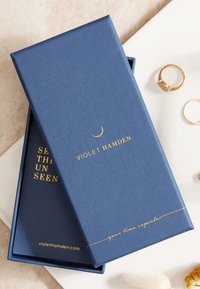 Boîte texturée bleu marine avec texte doré en relief. Sur le dessus se lit "VIOLET HAMDEN" et "votre capsule temporelle", avec un accent en forme de croissant de lune.