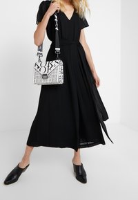 Vestido negro con escote en V y falda fluida, combinado con un bolso blanco con letras negras en negrita y un acabado texturizado. Mules negros.
