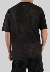 Hombre vistiendo camiseta negra de Reebok y pantalones cortos con patrones oscuros sutiles, logo rojo de Reebok visible cerca del cuello en la parte trasera.