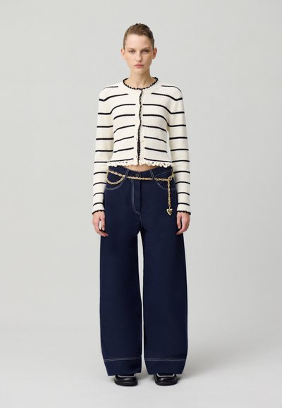 Cardigan en tricot rayé noir et blanc, avec un ourlet en dentelle. Assorti à un pantalon large bleu marine et une accent en chaîne dorée.