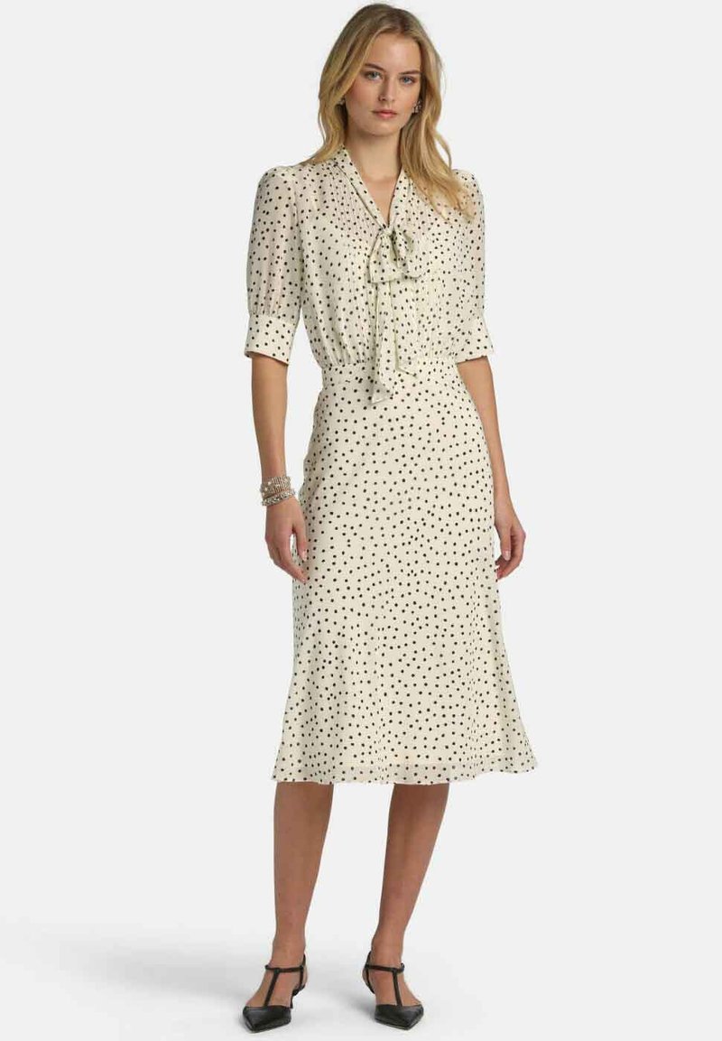 Krém színű pöttyös ruha rövid ujjakkal, nyakkendő részlettel a nyaknál, bővülő midi hosszúságban, és fekete pöttyökkel az egész anyagon.