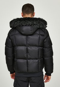 Svart dunjakke med stor faux fur-krage, quiltet design og ribbestrikkede cuffs. Materialet ser blankt og isolert ut.
