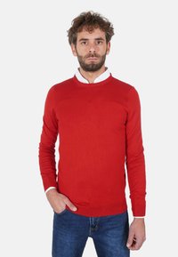Maglione rosso a maglia con maniche lunghe, collo tondo e polsini a coste. Indossato sopra una camicia bianca con colletto, abbinato a jeans blu.